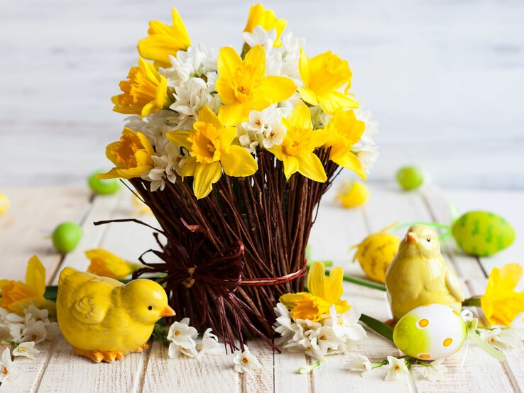  Ein frühlingshaftes Osterarrangement auf einem hellen Holztisch mit einem Strauß gelber Narzissen und weißen Blüten, die aus einem künstlichen Nest aus braunen Zweigen herausragen. Umgeben ist das Nest von dekorativen Elementen wie einem gelben Keramik-Küken, einem stehenden Vogel und verstreuten Ostereiern in verschiedenen Grüntönen, einige davon mit Schriftzug. Das Ensemble verkörpert die lebendige Frische des Frühlings und die Freude des Osterfests.