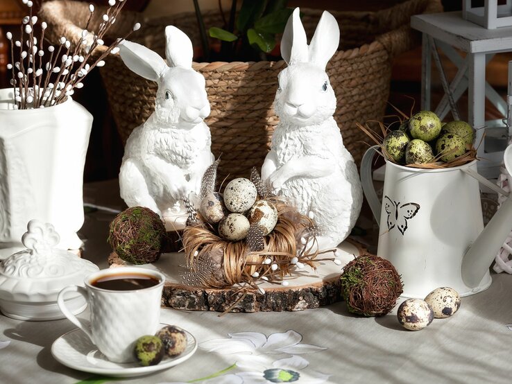 Das Bild zeigt eine festliche Osterdekoration, die sich durch eine natürliche und rustikale Ästhetik auszeichnet. Zwei weiße Keramikhasen stehen im Zentrum der Szene, flankiert von einem Arrangement aus Holzscheiben, die als Untersetzter dienen. Zwischen ihnen befindet sich ein Nest aus natürlichen Materialien, das sorgfältig mit dekorativen Eiern gefüllt ist, die mit einem gesprenkelten Muster versehen sind, was an Vogeleier erinnert. Ein weißer Krug mit einem Schmetterlingsmotiv beherbergt eine weitere Sammlung dieser Eier, was ein Gefühl von Fülle und Fruchtbarkeit vermittelt, was oft mit dem Osterfest in Verbindung gebracht wird.