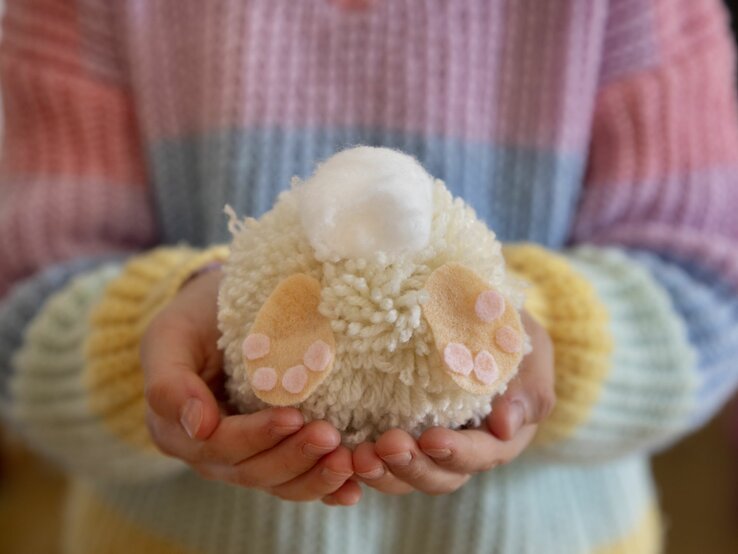  Das Bild zeigt ein handgefertigtes Osterdekorationsobjekt aus Wolle, das von zwei Händen gehalten wird. Die Hände sind um ein wolliges, rundes Objekt geschlossen, das scheinbar einen kleinen Osterhasen darstellt, komplett mit weißen und rosa farbenen Woll-Details, die die Pfoten und die Fußballen abbilden. Oben auf dem Objekt befindet sich eine kleinere weiße Wollkugel, die vermutlich den Schwanz des Hasen repräsentiert. Die Person trägt einen gestreiften Pullover in Pastelltönen, was eine weiche und warme Frühlingsstimmung vermittelt. Der Fokus auf das wollige Osterhasenobjekt in den sorgsam haltenden Händen unterstreicht die liebevolle und sorgfältige Herstellung von saisonaler Handwerkskunst.