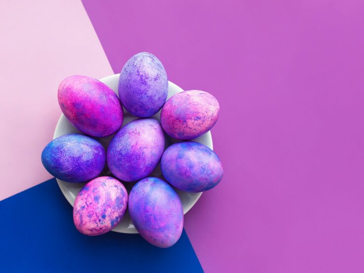 Das Bild zeigt eine Gruppe von Ostereiern, die mit einer marmorierten Technik in lebendigen Lila- und Blautönen gefärbt wurden, was auf die Verwendung von Rasierschaum als Färbemittel hinweisen könnte. Die Eier liegen auf einem weißen Teller, der wiederum auf einer zweifarbig geteilten Unterlage in Lila und Blau ruht, was das Farbthema der Eier aufgreift und einen starken visuellen Kontrast erzeugt. Diese Färbemethode erzeugt ein einzigartiges, wolkenartiges Muster auf den Eiern und ist eine kreative Alternative zu traditionellen Färbeverfahren für Ostereier.
