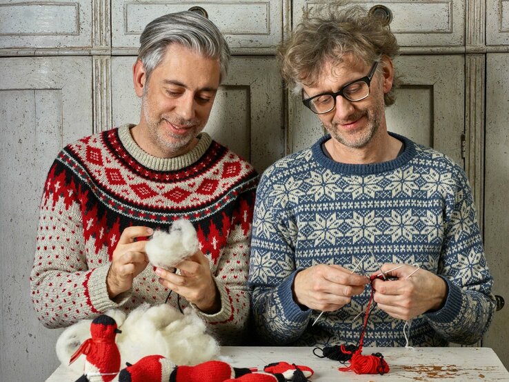 Zwei Männer, die gemeinsam Handarbeiten ausführen. Der Mann links im Bild trägt einen rot-schwarz-weißen Weihnachtspullover, der Mann rechts einen blau-weißen Pullover im skandinavischen Stil. Beide Männer haben graue Haare und scheinen in ihrer Handarbeit vertieft zu sein. Auf dem Tisch vor ihnen liegen fertige gestrickte Objekte. Sie sitzen vor einem Schrank mit antikem Aussehen | © Topp Verlag