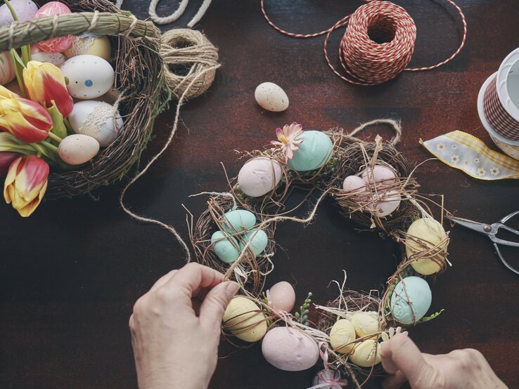 Eine Person bastelt auf einem dunklen Holztisch einen Osterkranz. Der Kranz ist mit verschiedenen pastellfarbenen Eiern verziert, die teilweise gemustert sind. Neben dem Kranz sind frische Tulpen und Bastelmaterialien wie Schnüre, Schere und zusätzliche Dekoelemente zu sehen.