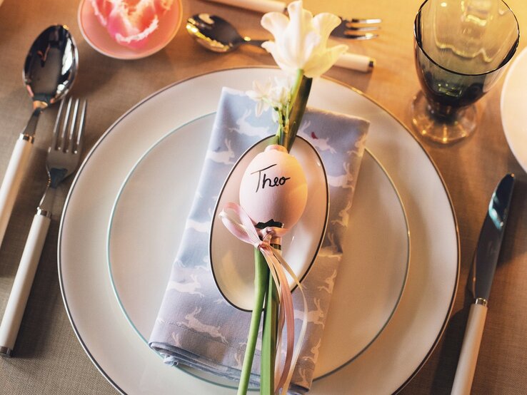 Festlich gedeckter Oster-Tisch. Auf dem Teller liegt eine Serviette, darauf ein Tischkärtchen aus Eierschale mit dem Namen "Theo". Darum ist ein Band gebunden, an dem eine einzelne Blume befestigt ist. | © Callwey Verlag