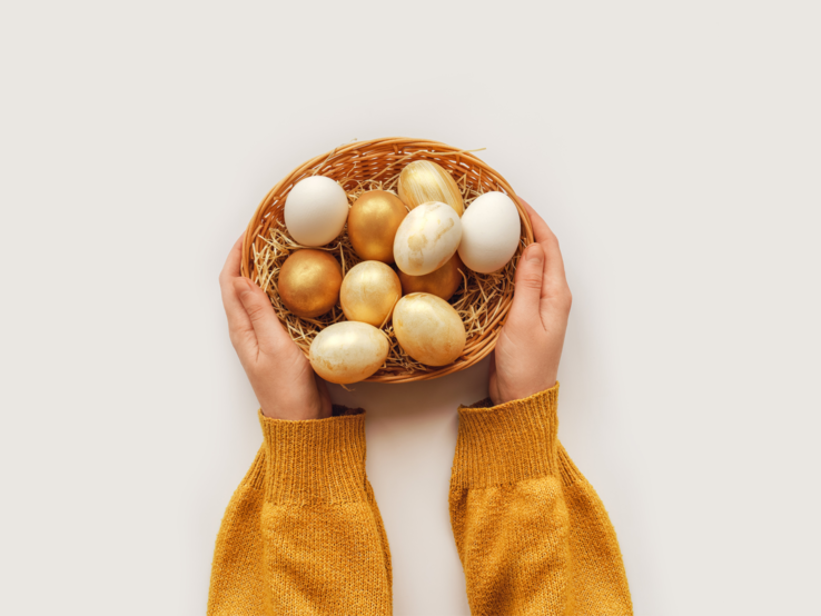  Das Bild zeigt die Hände einer Person, die einen Korb hält. In dem Korb befinden sich Heu und eine Sammlung von Eiern mit natürlicher weißer Schale und anderen, die goldfarben bemalt oder gefärbt sind. Der goldene Glanz verleiht den Eiern eine festliche und dekorative Erscheinung, die typisch für Osterdekorationen ist. Der Hintergrund ist einheitlich weiß, was die Farben der Eier und den natürlichen Ton des Korbes hervorhebt. Die Ärmel des Pullovers der Person sind strickig und von satter senfgelber Farbe, was warm und saisonal passend wirkt