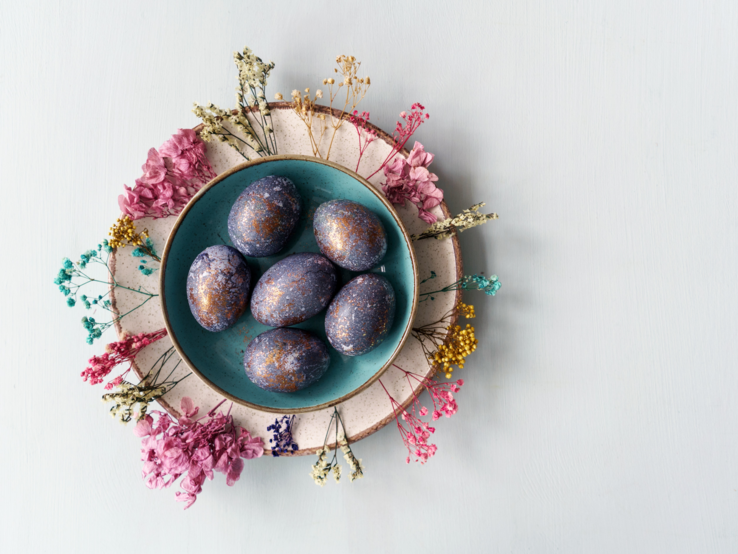 Das Bild zeigt einen liebevoll arrangierten Teller mit marmorierten Ostereiern, umgeben von getrockneten Blumen verschiedener Farben. Die Eier präsentieren sich in einer tiefblauen Farbe mit metallisch wirkenden, kupferfarbenen Sprenkeln, die eine kunstvolle und marmorierte Oberfläche erzeugen. Der Teller steht auf einem größeren, zweifarbigen Teller, der als Unterlage dient und das Arrangement abrundet. Der Hintergrund ist hell und neutral, was die Farbvielfalt der Eier und Blumen hervorhebt und eine ruhige, ästhetische Atmosphäre schafft.