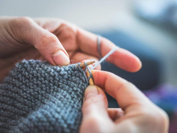 Detailaufnahme von Händen, die geschickt graue Wolle mit einer Stricknadel zu einem Schal verarbeiten.