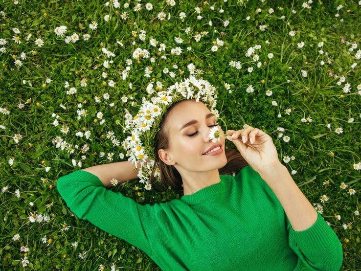 Junge Frau mit Gänseblümchenkranz und grünem Pullover liegt lächelnd auf einer Wiese und riecht an einer Blume.