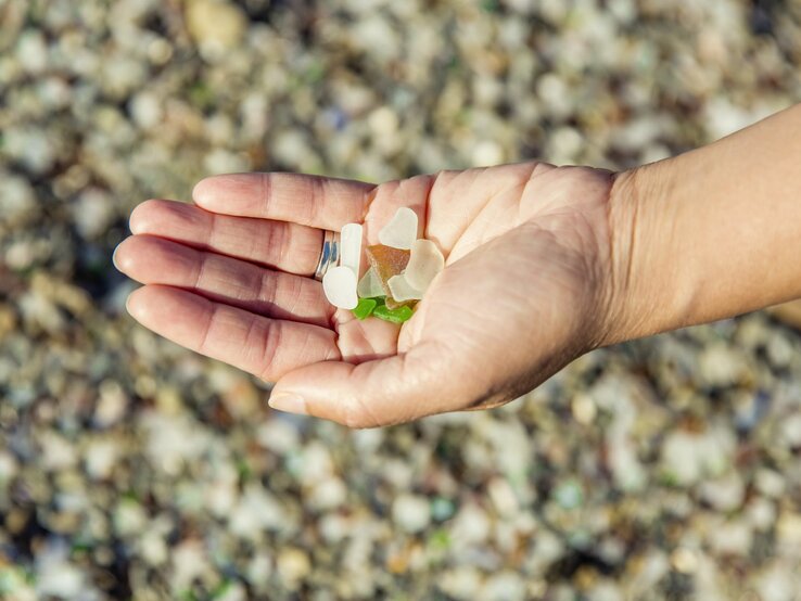 Eine Hand mit silbernem Ring hält bunte Glasstücke auf einem kieseligen Strand.