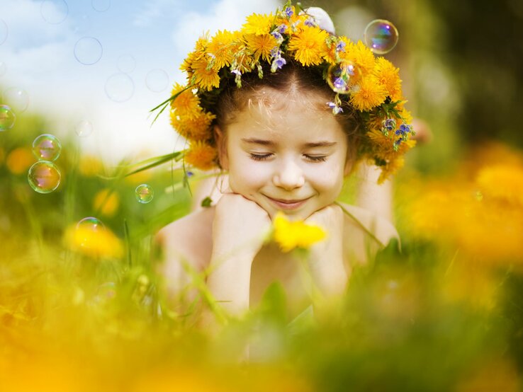 Ein junges Mädchen mit Blumenkranz im Haar lächelt mit geschlossenen Augen auf einer Wiese voller Löwenzahn und Seifenblasen.