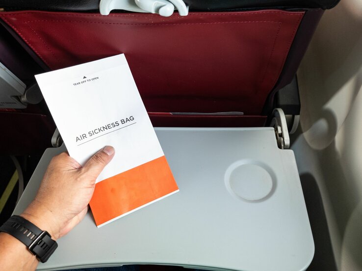 Basteln mit Spuckbeutel Eine Hand hält einen Spuckbeutel über einem heruntergeklappten Tablett in einem Flugzeug mit dunkelrotem Rücksitz.