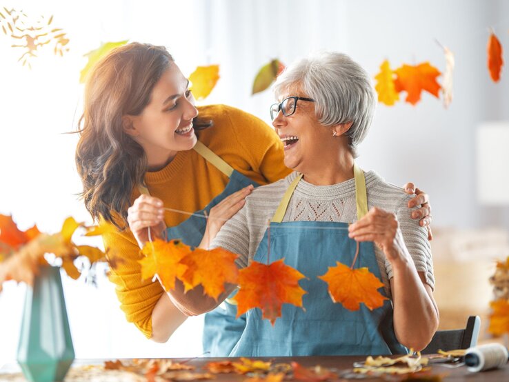 Herbstdeko für Wand basteln, Wanddeko Herbst, Wanddeko Herbst selbst machen, DIY-Wanddeko Herbst Eine lachende ältere Frau mit grauen Haaren bastelt mit einer jungen Frau mit braunen Haaren an Herbstlaubketten in einem sonnigen Raum.