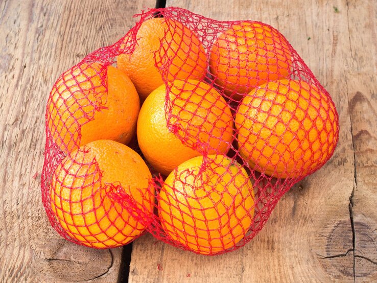 Ein rotes Netz mit sieben frischen Orangen liegt auf rustikalem Holzuntergrund.