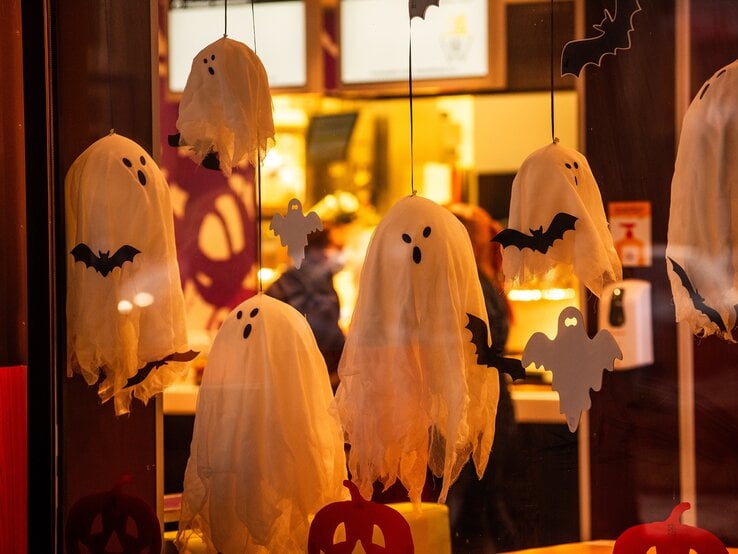 Geister basteln  Weiße Geister und schwarze Fledermäuse aus Stoff hängen als Halloween-Dekoration hinter einer erleuchteten Glasscheibe.