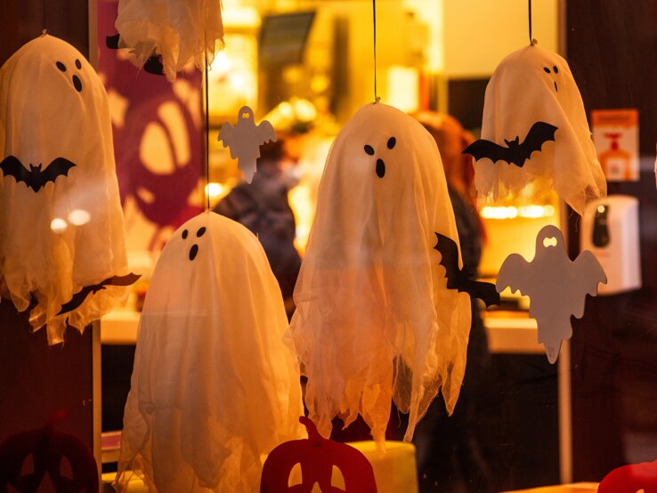 Geister basteln  Weiße Geister und schwarze Fledermäuse aus Stoff hängen als Halloween-Dekoration hinter einer erleuchteten Glasscheibe.