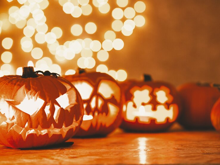 Last-minute-Halloween-Deko, Last-minute-Halloween-Deko basteln, DIY Last-minute-Halloween-Deko, Bastelideen Last-minute-Halloween-Deko Leuchtende Kürbislaternen mit gruseligen Fratzenschnitzereien stehen auf einem Holztisch vor unscharfen Lichterketten im Hintergrund.