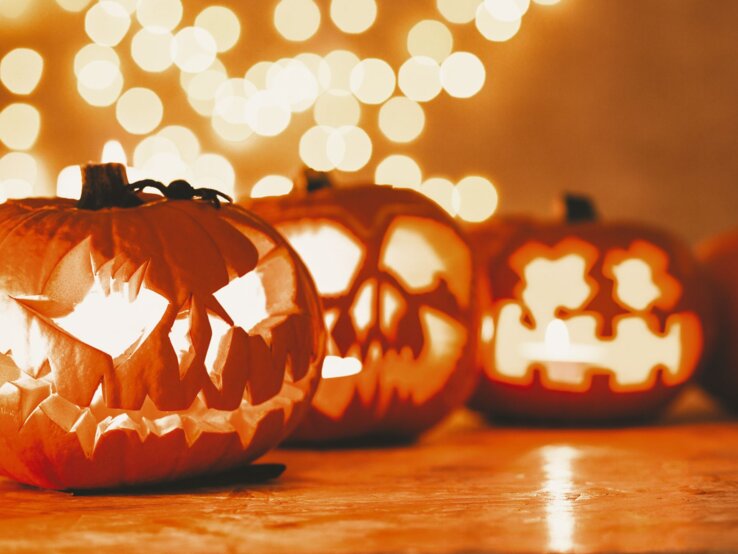 Last-minute-Halloween-Deko, Last-minute-Halloween-Deko basteln, DIY Last-minute-Halloween-Deko, Bastelideen Last-minute-Halloween-Deko Leuchtende Kürbislaternen mit gruseligen Fratzenschnitzereien stehen auf einem Holztisch vor unscharfen Lichterketten im Hintergrund.