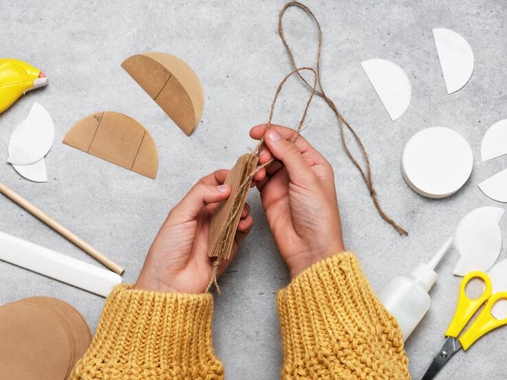 Hände in gelbem Strickpullover binden mit Juteschnur Pappscheiben zusammen, umgeben von Bastelmaterial, darunter Kaffeefilter, auf grauem Tisch.