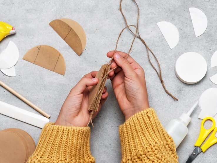 Hände in gelbem Strickpullover binden mit Juteschnur Pappscheiben zusammen, umgeben von Bastelmaterial, darunter Kaffeefilter, auf grauem Tisch.