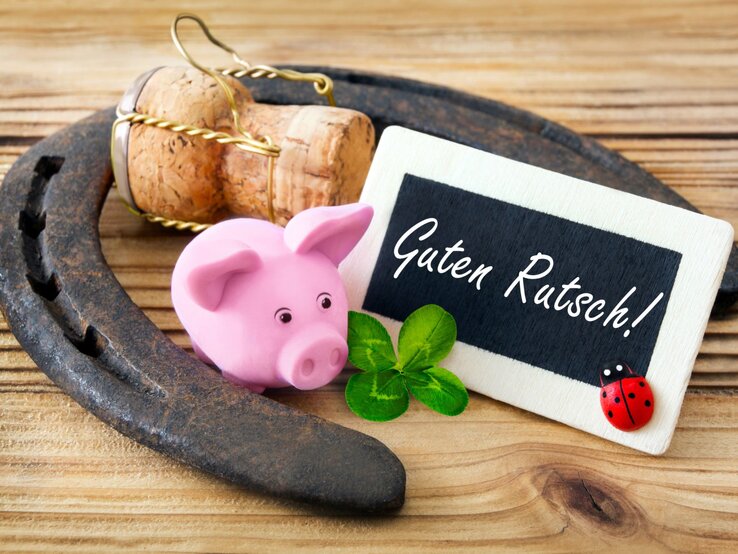 Glücksschwein basteln, DIY-Glücksschwein, Silvester-Glücksschwein, Glücksbringer Schwein Ein rosafarbenes Glücksschwein, ein Kleeblatt, ein Hufeisen und eine Holztafel mit der Aufschrift „Guten Rutsch!“ liegen auf hellem Holz.
