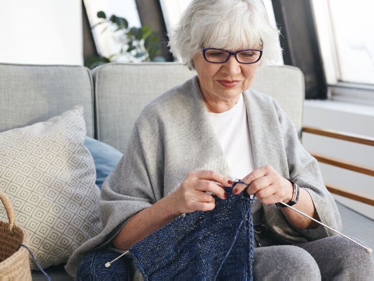 Ältere Frau mit grauen Haaren und Brille strickt konzentriert ein dunkelblaues Tuch auf einem grauen Sofa.