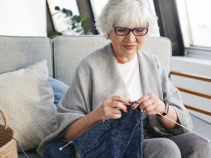 Ältere Frau mit grauen Haaren und Brille strickt konzentriert ein dunkelblaues Tuch auf einem grauen Sofa.