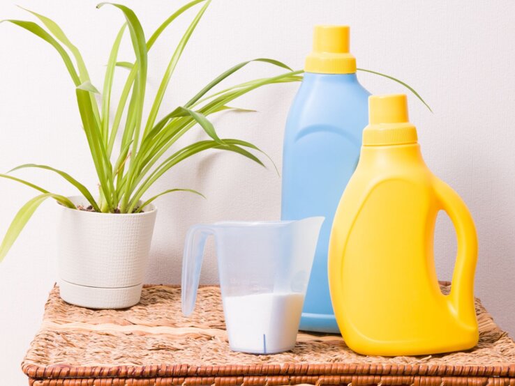 Waschmittelflasche Upcycling Zwei bunte Waschmittelflaschen in Gelb und Blau stehen neben einem Messbecher mit Pulver und einer Zimmerpflanze auf einem Flechtkorb.