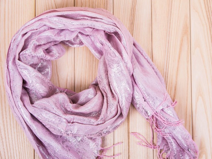Ein glänzender, rosa Schal mit feinen Fransen liegt locker zu einem Kreis auf hellen Holzlatten drapiert.
