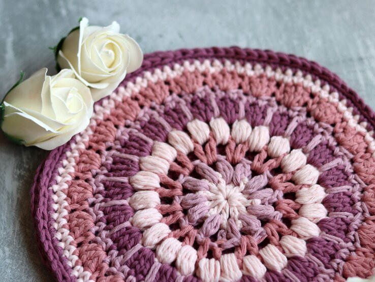 Topfuntersetzer basteln, DIY-Topfuntersetzer, Topfuntersetzer selbst machen, Topfuntersetzer selbst basteln Ein Topfuntersetzer als handgefertigte Häkelarbeit mit kreisförmigem Farbverlauf in Rosa und Lila, ergänzt durch zwei edle weiße Rosen auf Betonhintergrund.