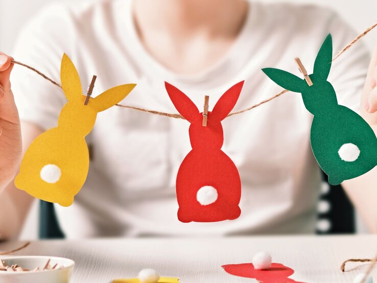 Eine Person hält eine Oster-Girlande mit bunten Papierhasen in Gelb, Rot und Grün, verziert mit weißen Bommel-Schwänzchen.