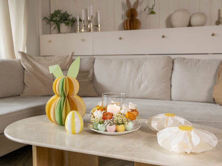 Farbiger Wabenhase aus Papier mit grünen Ohren steht neben Deko-Ei und Osterkranz aus Blumen und Kerzen auf einem Couchtisch.