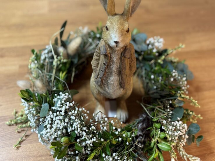Auf Holzboden steht ein sitzender Hasenfigur mit Weste in einem Osterkranz aus Schleierkraut, Eukalyptus, Getreide und Grünzweigen.