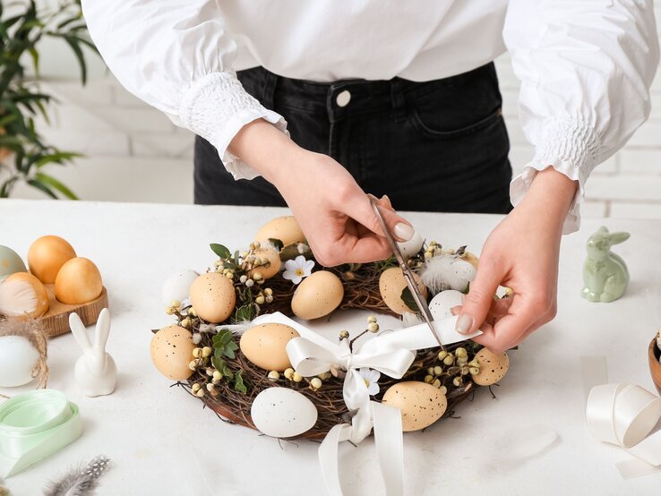 Junge Frau mit weißer Bluse bindet ein weißes Band an einen Osterkranz aus Zweigen, Eiern, Blüten und Beeren auf weißem Tisch.