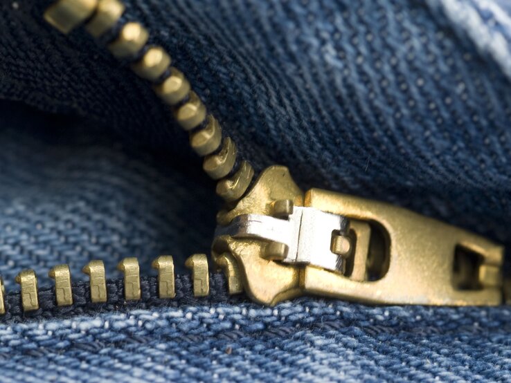 Goldener Reißverschluss in Nahaufnahme auf blauem Jeansstoff, leicht geöffnet, mit scharf sichtbarer Struktur des Gewebes.