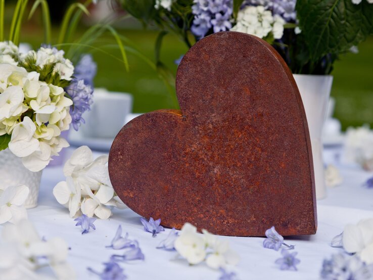 Rostiges Herz aus Metall steht dekorativ zwischen weißen und violetten Blüten auf festlich gedecktem Tisch im Garten.