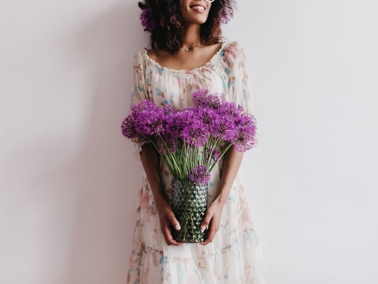 Junge Frau mit lockigem Haar und geblümtem Kleid hält lächelnd eine Vase mit violett blühendem Allium (Zierlauch) vor hellem Hintergrund.