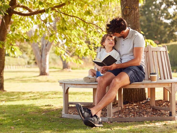 Baumbank DIY Junger Vater liest seinem lachenden Sohn auf einer Baumbank unter grünem Baum liebevoll ein Buch vor.