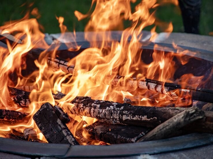 Loderndes Feuer in einer runden DIY-Feuerschale mit glühenden Holzscheiten vor unscharfem grünem Hintergrund am Abend.