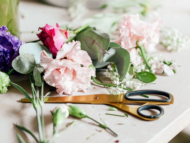 Frisch geschnittene Blumen in Pastellfarben und eine offene Schere deuten auf kreative Floristikarbeit auf hellem Untergrund.