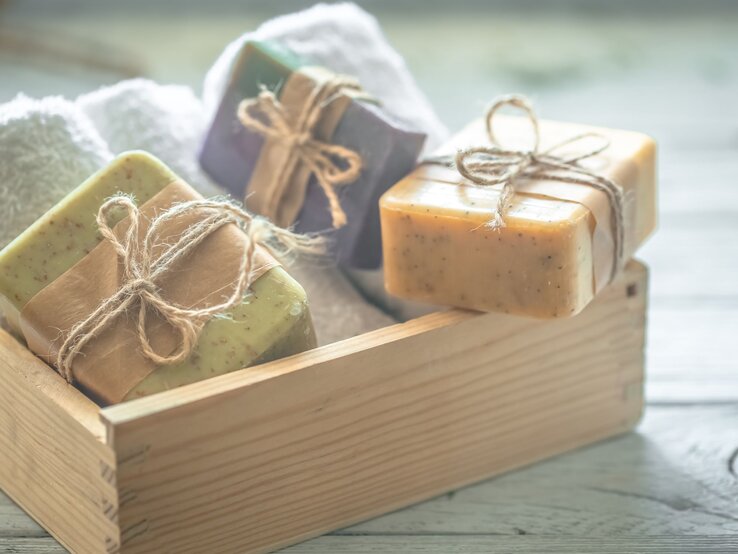 Drei DIY-Shampoos und handgemachte Seifenstücke mit Bastschleifen liegen in einer Holzkiste neben weißen, weichen Handtüchern.