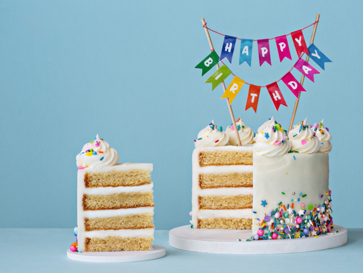 Weiß glasierte Geburtstagstorte mit bunten Streuseln und selbst gebastelter "Happy Birthday"-Cake Topper vor hellblauem Hintergrund.