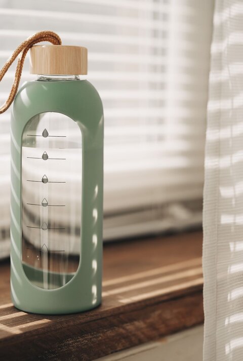 Moderne Glasflasche mit DIY-Wasser-Tracker leuchtet im warmen Licht vor halb geöffneten Jalousien.