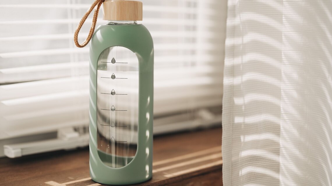 Moderne Glasflasche mit DIY-Wasser-Tracker leuchtet im warmen Licht vor halb geöffneten Jalousien.