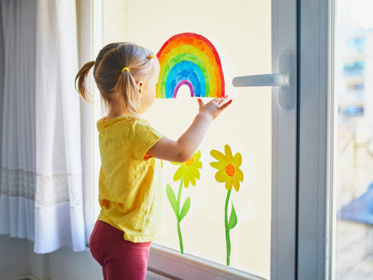 Kleines Mädchen mit blonden Zöpfen betrachtet fröhlich einen bunten Regenbogen am Fenster neben gemalten Sonnenblumen.