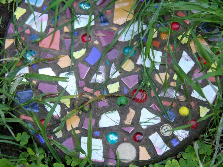 DIY Trittstein Garten Bunter Trittstein mit Glasscherben, Knopf und Murmeln liegt von hohem Gras umgeben auf Erde im Garten.