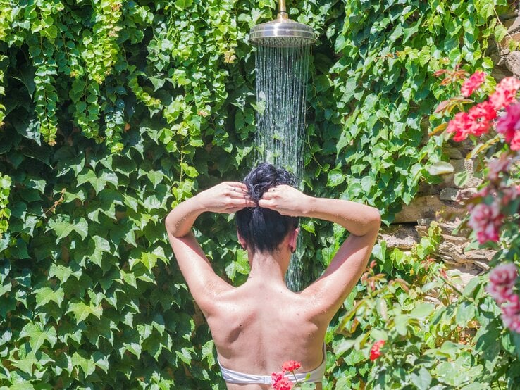 Frau mit dunklem Haar duscht im Freien vor einer mit Efeu bewachsenen Steinwand unter Sonnenlicht.