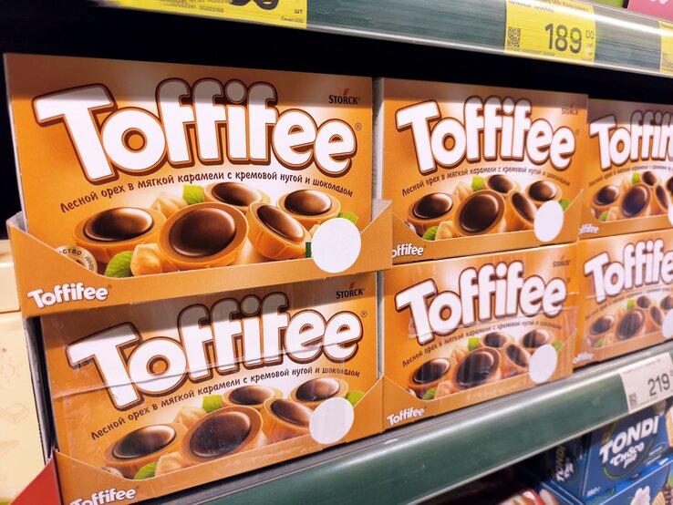 Nahaufnahme eines Supermarktregals mit orangefarbenen Toffifee-Schachteln und Preisschildern in kyrillischer Schrift.