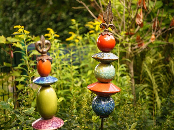 Zwei bunte Gartenstecker aus glasierten Keramikelementen mit metallischer Spitze stehen dekorativ im grünen Sommergarten.