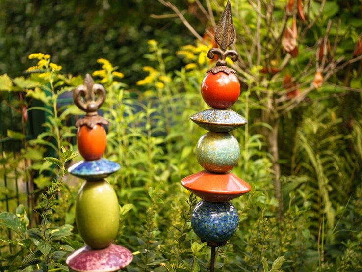 Zwei bunte Gartenstecker aus glasierten Keramikelementen mit metallischer Spitze stehen dekorativ im grünen Sommergarten.