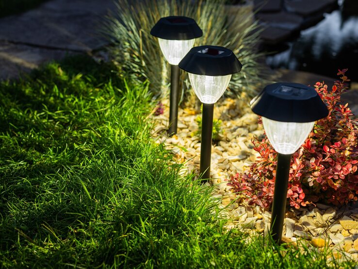 Solarlampe Garten DIY Basteln Drei leuchtende Solarlampen säumen einen Gartenweg mit Zierkies, roten Sträuchern und frisch gemähtem Rasen.