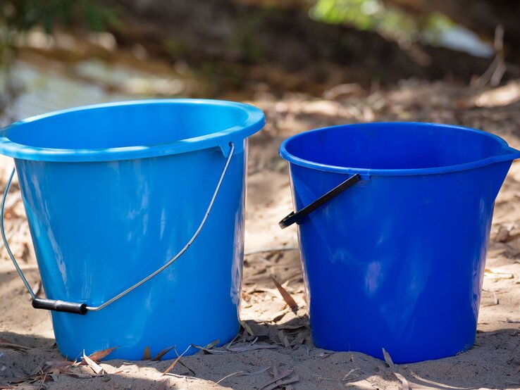 Plastikeimer in verschiedenen Blautönen, auf staubigem Untergrund mit verstreuten Blättern und leicht sonnigem Hintergrund.